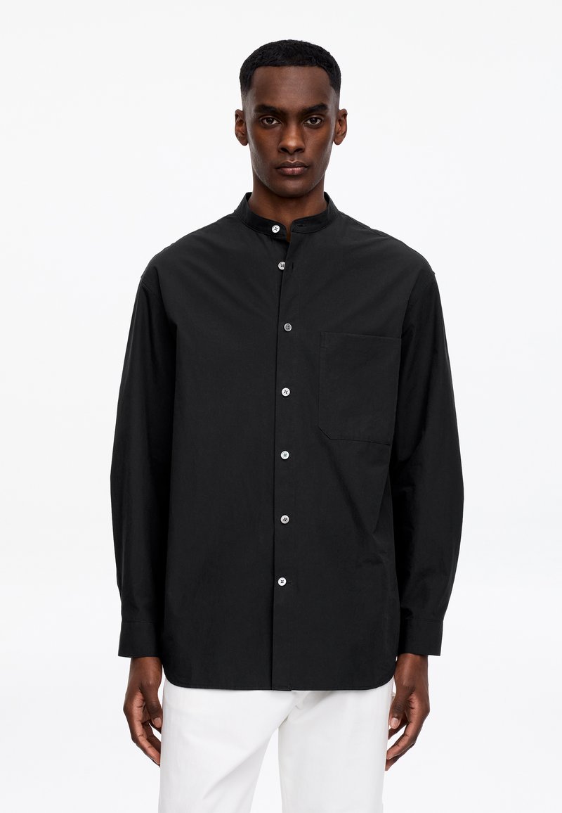 Homme portant une chemise noire à manches longues avec des boutons blancs et une poche avant, associée à un pantalon blanc, sur un fond blanc uni.