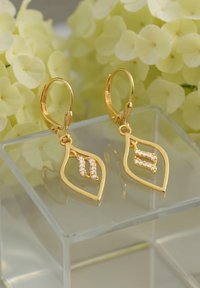 Orecchini a forma di foglia d'oro con pietre bianche scintillanti, caratterizzati da un design dettagliato e chiusure a leva. Inquadrati su uno sfondo floreale chiaro.