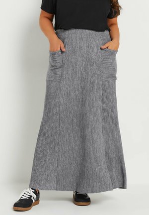 Vrouw draagt een lange, geribbelde grijze rok met grote voorzakken en zwarte sneakers met witte veters, staand tegen een effen achtergrond.