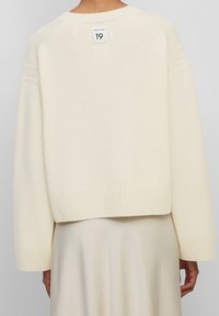 Pull tricoté couleur crème avec un design court, des poignets et un ourlet côtelés, comportant une étiquette d'entretien visible au niveau de l'encolure arrière.