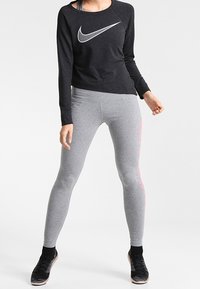 Maglietta nera a maniche lunghe con logo Nike bianco, abbinata a leggings grigi con accenti rosa. Il modello indossa scarpe da ginnastica nere.