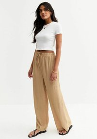 Camiseta blanca recortada y pantalones anchos beige sueltos con un acabado texturizado, que presentan una cintura con cordón y chanclas negras.
