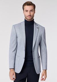 Blazer grigio chiaro in tessuto strutturato, con rever a punta, due tasche anteriori, dettagli blu e un singolo emblema rosso vicino alla spalla sinistra.