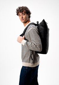 Zaino nero a ribalta con finitura lucida, spallacci regolabili e tasca esterna con cerniera, indossato da una persona con un maglione a righe e jeans.