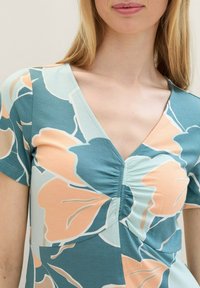 Frau trägt ein kurzärmliges V-Ausschnitt-Oberteil mit gerafftem Mittelteil und großem Blumenmuster in Türkis-, Minz- und Pfirsichtönen.