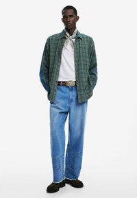 Conjunto casual que incluye una camisa de cuadros verdes, una camiseta blanca, jeans anchos azules con parches y zapatos negros. Incluye una bufanda con estampado y un cinturón.