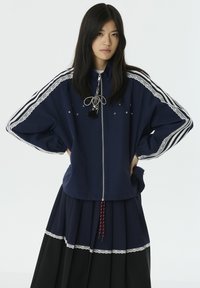 Marineblauwe zip-up jas met witte strepen en kantaccenten, versierd met sterren en een hangend sieraad. Gelaagd over een geplooide zwarte rok.