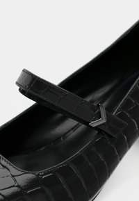 Chaussure Mary Jane en cuir noir avec un motif en relief de crocodile, dotée d'une sangle plate et d'un accent métallique là où la sangle rencontre la chaussure.