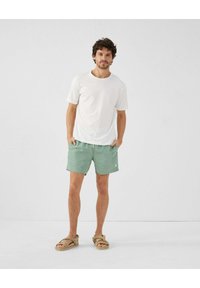 Witte t-shirt met korte mouwen en groene shorts met zijzakken. Model draagt beige sandalen. Eenvoudige, relaxte pasvorm en effen kleuren.
