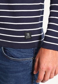 Camisa de rayas azul marino y blanca de manga larga en suave tela de punto, con una etiqueta negra que dice "TTD ORIGINAL CLOTHING" cerca del dobladillo.