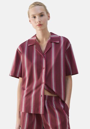 Kvinde iført en kortærmet skjorte med lodrette pink og burgunder striber samt matchende shorts, stående med den ene hånd i lommen.