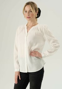 Bílá blůza s dlouhým rukávem a knoflíkovým zapínáním, měkká tkanina, nařasené ramena a zakřivený lem, spárovaná s černými džíny.