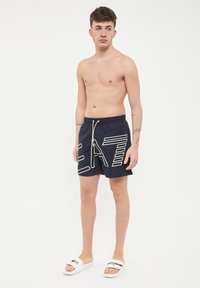 Marinblå simshorts med vit grafisk text, elastisk midje och snören. Modell som bär vita sandaler, stående mot en neutral bakgrund.