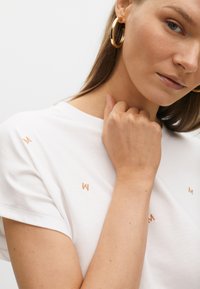 T-shirt en coton blanc avec de petites lettres brodées "M" en or rose. Manches courtes et coupe décontractée, porté avec un geste de la main visible.