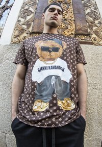 Bruine graphic T-shirt met een beer in een zonnebril en een wit logo-shirt, aangevuld met gouden sneakers en een gemusterde achtergrond.