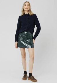 Blouse marine à manches longues avec détails en volants et fermeture à boutons, associée à une mini-jupe vert brillant. Chaussures à lacets marron et chaussettes noires.