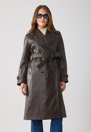 Bruine leren trenchcoat met dubbelzijdige knopen, ceintuur in de taille en brede revers; model draagt blauwe zonnebril en denim broek.