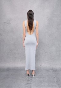 Vestido maxi gris sin mangas con un profundo escote en la espalda, hecho de un tejido elástico, con una silueta ajustada. Lleva sandalias de tacón blancas.