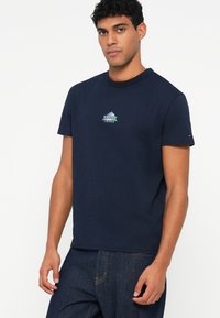 Ναυτικό μπλε βαμβακερή μπλούζα T-shirt με κανονική λαιμόκοψη, που διαθέτει ένα μικρό λογότυπο βουνού σε πράσινο και λευκό στο στήθος, και κοντά μανίκια.