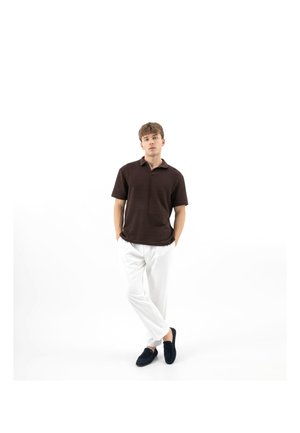 Junger Mann steht mit den Händen in den Taschen, trägt ein dunkelbraunes, strukturiertes Polohemd mit kurzen Ärmeln, weiße Hosen und dunkle Slipper auf weißem Hintergrund.