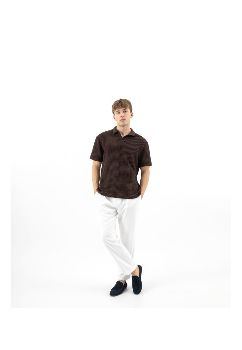 Giovane uomo in piedi con le mani nelle tasche, che indossa una polo a maniche corte marrone scuro con trama, pantaloni bianchi e scarpe slip-on scure su sfondo bianco.