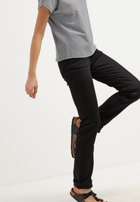 Pantaloni neri slim-fit realizzati in tessuto resistente, abbinati a una maglietta a righe blu navy e bianca con maniche corte e sandali neri con cinturini.