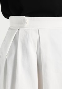 Jupe plissée blanche avec une texture lisse et un détail de bouton à la taille. Le tissu est robuste et léger, offrant une chute structurée.