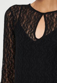 Haut en dentelle noire à manches longues avec motif floral, doté d'un décolleté en forme de goutte et de manches semi-transparentes sur la peau.