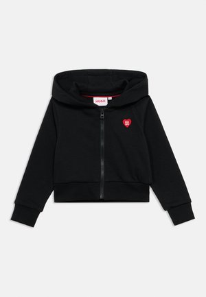 Sudadera corta negra con cierre de cremallera, con un logo de corazón rojo en el pecho izquierdo, puños acanalados y textura de tela suave.