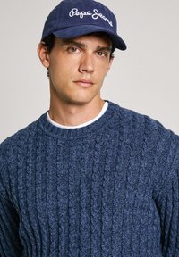 Temno moder pleteni pulover z vzorcem združenih pletenic in okroglim izrezom, v kombinaciji z modro kapo, na kateri je v belih črkah izšito "Pepe Jeans".