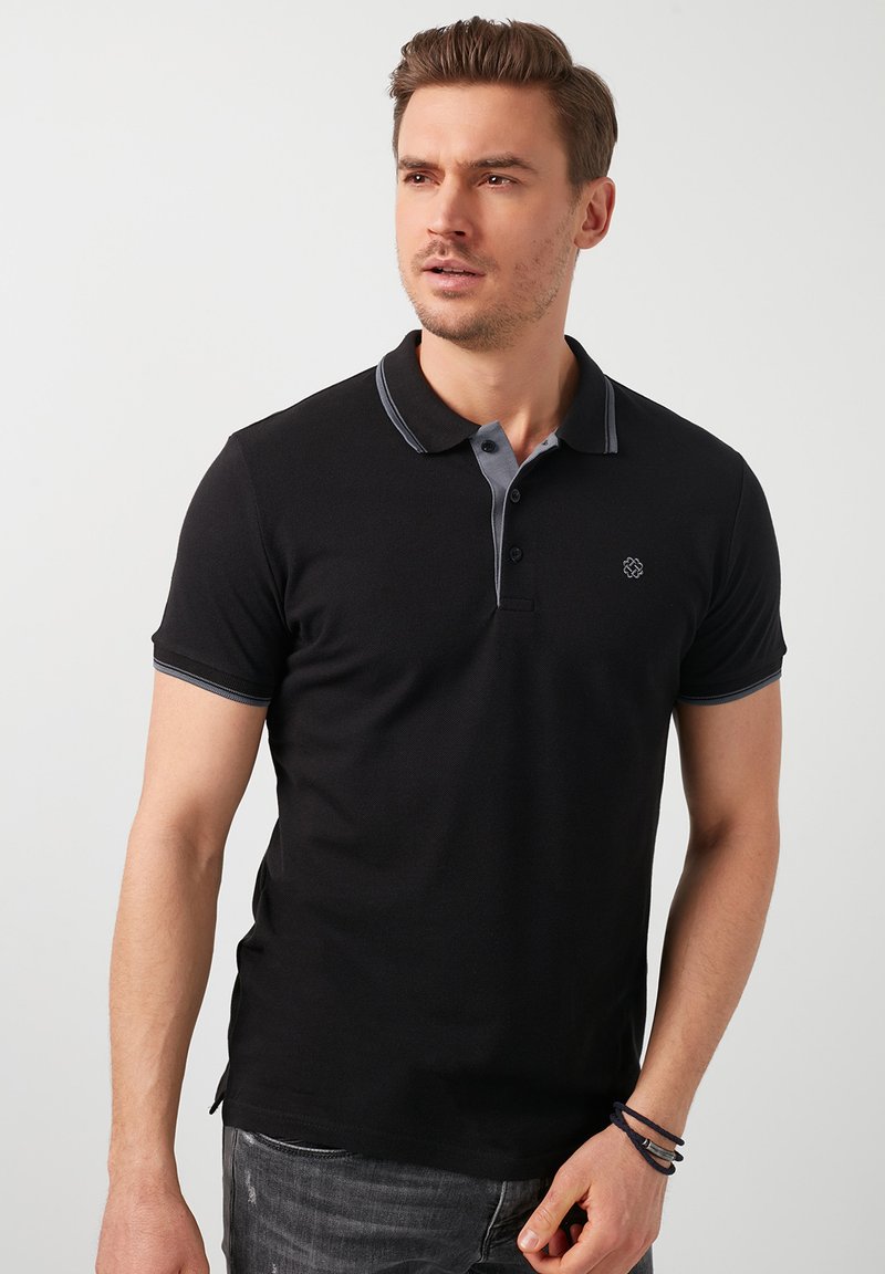 Polo shirt nera con colletto e pattina grigi, dotata di tre bottoni. Maniche corte con dettagli grigi. Semplice logo ricamato sul petto.