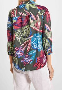 Camisa con estampado floral que presenta verdes, rojos y azules vibrantes sobre un fondo oscuro. Tela suave, cuello abotonado y mangas tres cuartos.