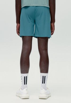 Personne portant un short de sport bleu sarcelle, des chaussettes blanches à rayures noires et des baskets blanches, debout de dos sur un fond uni.