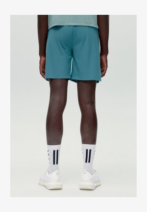 Personne portant un short de sport bleu sarcelle, des chaussettes blanches à rayures noires et des baskets blanches, debout de dos sur un fond uni.