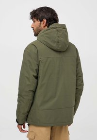 Giacca a cappuccio verde oliva con una texture liscia e opaca. Caratterizzata da una linea dritta e da un cappuccio regolabile, con tasche laterali con zip.