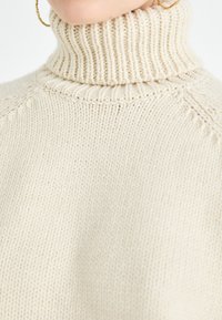 Beige knitted turtleneck sweater featuring a ribbed design and textured stitch patterns, with a relaxed fit and detailed neckline.