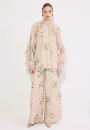 Femme portant un chemisier à volants fleuri pêche avec des manches larges et un pantalon large assorti sur un fond blanc uni.