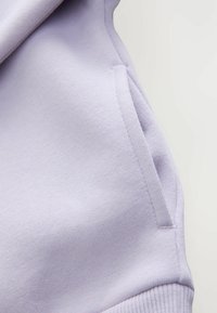 Felpa lilla caratterizzata da una texture morbida, polsini a costine e un dettaglio con tasca laterale, enfatizzando il comfort e lo stile casual.