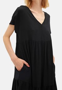 Žena v volných černých šatech s výstřihem ve tvaru V a krátkými rukávy, s rukama v postranních kapsách, na světlém jednolitém pozadí.