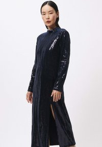 Marineblauwe pailletten jurk met lange mouwen en een hoge halslijn. Heeft een split tot boven de dij voor extra detail, wat zorgt voor een slanke silhouet.