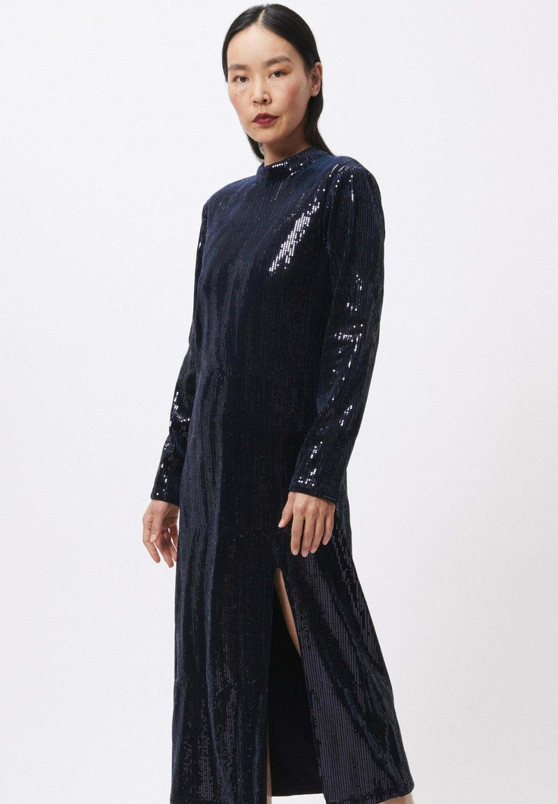 Marineblauwe pailletten jurk met lange mouwen en een hoge halslijn. Heeft een split tot boven de dij voor extra detail, wat zorgt voor een slanke silhouet.