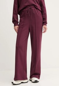 Bordové tepláky s uvoľneným strihom, elastickým pásom, šnúrkou, bočnými švami a textúrovanou tkaninou; v kombinácii s bielymi športovými teniskami.