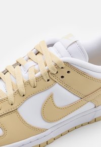 Vit och ljusbeige sneaker med ovandel i läder och tyg, runda snören och en framträdande Nike-logotyp i beige på sidan.