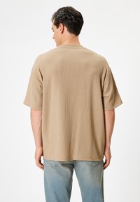 Kortærmet, beige tekstureret skjorte med en afslappet pasform, der har en rund halsudskæring og synlige sømdetaljer bagpå. Båret med lyseblå jeans.
