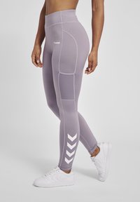 Lilla leggings med høj talje, strukturerede paneler og hvid chevron-detaljer. Stylet med hvide sneakers på en neutral baggrund.