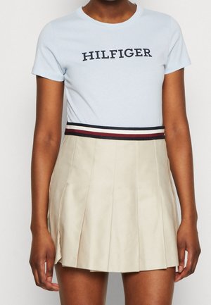 Osoba w jasnoniebieskiej koszuli z krótkim rękawem z napisem "HILFIGER" oraz w beżowej spódnicy plisowanej z paskiem w paski w kolorze granatowym, białym i czerwonym.