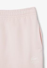 Pantaloni della tuta rosa chiaro con vita elastica arricciata, tasche laterali e logo Lacoste bianco sulla parte inferiore della gamba sinistra. Tessuto morbido.