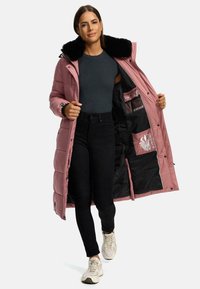 Palton pufos lung de culoare roz, cu guler din blană sintetică neagră, având buzunare interioare, închidere cu fermoar și căptușeală neagră. Modelul poartă pantaloni negri și adidași.