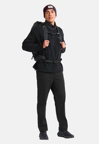 Jeune homme portant un équipement de plein air noir et un bonnet violet, se tenant avec les mains tenant les sangles d'un sac à dos devant un fond uni.