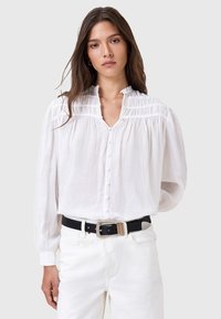 Blusa bianca con bottoni, colletto arricciato e dettagli plissettati. Maniche lunghe, vestibilità rilassata, tessuto leggero. Abbinata con una cintura nera e pantaloni bianchi.